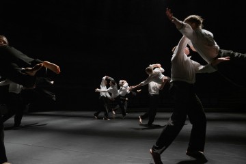 Ryhmä valkoisiin toppiin ja mustiin housuihin pukeutuneita tanssijoita tekee dynaamisia nostoja ja liikkeitä hämärästi valaistulla näyttämöllä luoden dramaattisia varjoja ja liikkeen tunnetta.