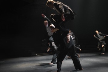 Neljä tummiin vaatteisiin pukeutunutta tanssijaa esiintyy hämärästi valaistulla lavalla. Yksi tanssija nostetaan korkealle muiden yläpuolelle, ja muut liikkuvat energisesti luoden dynaamista liikettä ja dramaattisia varjoja.