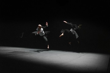 Kaksi tummiin pukuihin pukeutunutta ihmistä hyppää ilmaan hämärästi valaistulla näyttämöllä, ja heidät on kuvattu kesken liikkeen mustaa taustaa vasten, mikä viittaa dramaattiseen tanssiin tai esitykseen.