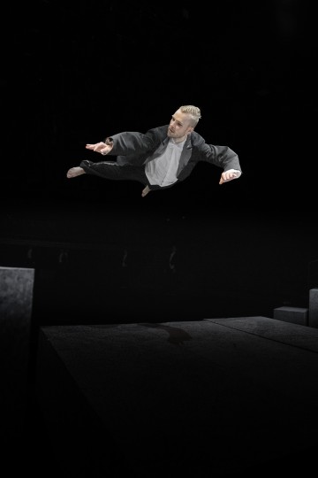 Pukuun pukeutunut henkilö on kuvattu keskellä ilmaa tummaa taustaa vasten, ja hän näyttää hyppäävän tai lentävän vaakatasossa suurten betoniharkkojen yläpuolella.