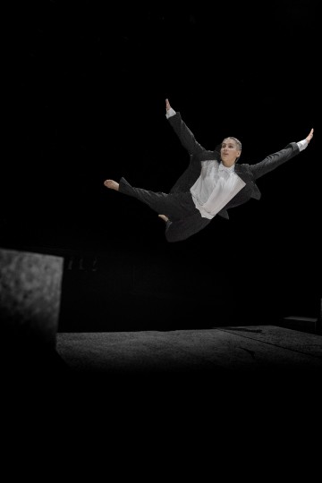 Pukuun pukeutunut henkilö tekee vaikuttavan ilmassa tapahtuvan hajautetun hypyn kädet ojennettuina, ja taustalla on tumma tausta, jossa on kivimäisiä alustoja.