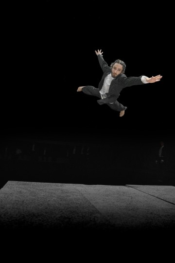 Paljasjalkainen, puku päällä oleva henkilö on kuvattu ilmassa dramaattisessa hypyssä kädet ja jalat levällään tummalla taustalla valaistun näyttämön yläpuolella.