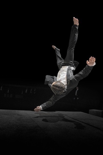 Pukuun pukeutunut henkilö on kuvattu keskellä ilmaa, ylösalaisin ja kädet ojennettuina, kun hän tekee akrobaattisen voltin tummaa taustaa vasten.