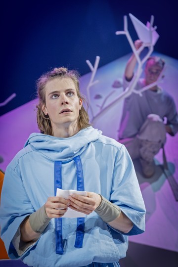 Nuori henkilö, jolla on löysä sininen vaate, pitää paperia kädessään ja katsoo mietteliäästi ylöspäin. Taustalla toinen hahmo istuu valkoisten oksien äärellä taittelemassa paperiveneitä värikkään, pehmeästi valaistun lavastuksen edessä.