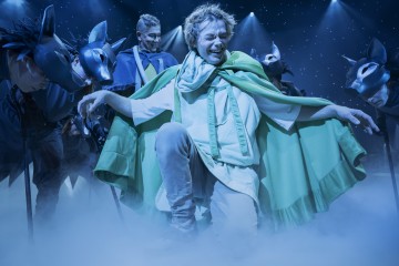 Vihreään viittaan pukeutunut esiintyjä polvistuu hymyillen keskellä näyttämöä, ja häntä ympäröivät tummiin pukuihin ja susinaamareihin pukeutuneet näyttelijät, ja sininen valaistus ja sumu luovat unenomaisen teatteritunnelman.