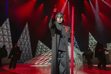 Tummaan kaapuun ja kypärään pukeutunut henkilö pitelee korkeaa punaista sauvaa ja osoittaa ylöspäin punaisella valaistulla näyttämöllä, jossa on geometrisia muotoja. Taustalla seisoo muita samalla tavalla pukeutuneita hahmoja, joiden rinnassa on punainen X-kirjain.