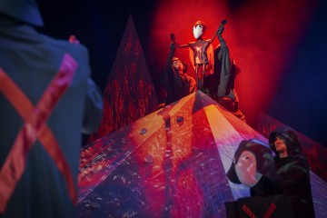 Dramaattisessa näyttämökohtauksessa on valkoisiin naamioihin verhoutuneita esiintyjiä hehkuvan, kulmikkaan vuoren ympärillä. Yksi hahmo seisoo voitokkaasti vuoren huipulla ja pitää kädessään punaisessa valossa kylpevää nukkea tai naamiota.