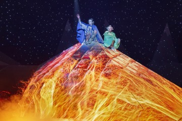 Kaksi värikkäisiin pukuihin pukeutunutta näyttelijää istuu näyttämötulivuoren huipulla, jossa on kirkkaita, tulisia valotehosteita ja savua, ja taustalla on tähtiä, jotka luovat dramaattisen kohtauksen.