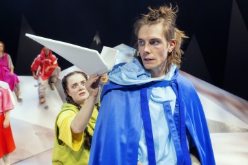 Siniseen viittaan pukeutunut henkilö katsoo intensiivisesti eteenpäin, kun taas toinen keltaisiin pukeutunut henkilö pitää suurta paperilentokonetta olkapäänsä lähellä. Muut värikkäisiin asuihin pukeutuneet istuvat taustalla valkoisella, kulmikkaalla näyttämöllä.