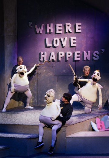 Neljä esiintyjää käyttää suuria vauvanukkeja lavalla seinän edessä, jossa lukee isoin kirjaimin WHERE LOVE HAPPENS. Kohtaus on värikäs ja teatraalinen, ja siinä on dramaattinen valaistus ja ilmeikkäät nukkekasvot.