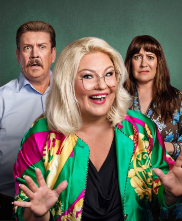 Hymyilevä vaalea nainen, jolla on silmälasit ja värikäs takki, seisoo edessä ja elehtii käsillään, kun taas hänen takanaan vihreällä taustalla seisovat yllättynyt mies ja huolestunut nainen.