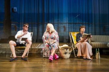 Kolme aikuista istuu lavasteessa, joka on suunniteltu näyttämään rannalta, ja taustalla on sininen meri. Yksi aurinkolasipäinen mies elehtii puhuessaan, hymyilevä nainen istuu mekossaan ja toinen nainen lukee kirjaa. Rantakassit ovat heidän jaloissaan.