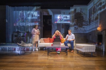 Kolme ihmistä on modernissa olohuoneessa kannettavan tietokoneen kanssa; yksi istuu vaaleanpunaisella sohvalla ja elehtii vilkkaasti, kun muut seisovat tai istuvat lähellä. Taustalle heijastetuilla näytöillä näkyy useita kasvoja, jotka viittaavat videopuheluun.