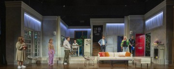 Seitsemän näyttelijää seisoo kirkkaasti valaistulla näyttämöllä, joka on moderni olohuone, jossa on harmaat seinät, suuret ikkunat, portaat ja punainen ovi. Ryhmä näyttää keskustelevan, mikä luo vilkkaan kohtauksen.