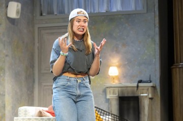 Henkilö, jolla on takapuolinen lippis, harmaa crop-toppi ja farkut, seisoo sisätiloissa vihainen ilme kasvoillaan ja turhautuneena kädet koholla, ja näyttää esiintyvän lavalla.