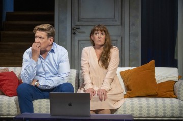 Mies ja nainen istuvat jännittyneinä sohvalla ja näyttävät huolestuneilta. Mies koskettaa kasvojaan mietteliäänä, kun taas nainen kurottaa kätensä syliinsä. Kannettava tietokone on avoinna heidän edessään pöydällä. Tyynyt ovat heidän vieressään.