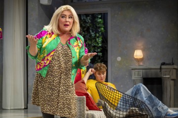 Nainen, jolla on vaaleat hiukset, silmälasit ja värikäs takki, elehtii seisaaltaan avoimin käsin. Hänen takanaan keltaisiin pukeutunut nuori istuu sohvalla ja katsoo eteenpäin. Ympäristö näyttää olevan olohuone.