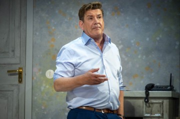 Vaaleansiniseen paitaan pukeutunut mies seisoo sisätiloissa ja osoittaa oikealla kädellään. Hän näyttää puhuvan. Hänen takanaan on suljettu ovi, pilkullinen seinä, kirjoituspöytä ja puhelin.