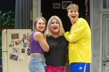 Kolme ihmistä seisoo yhdessä keittiössä hymyillen ja halaten. Nuori nainen violetissa topissa, vaaleahiuksinen ja silmälasipäinen henkilö mustissa silmälaseissa ja nuori mies keltaisessa paidassa näyttävät iloisilta ja innostuneilta.