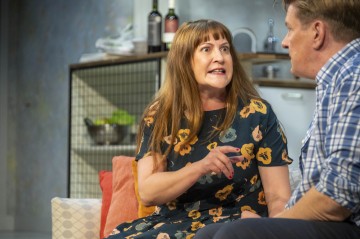 Nainen, jolla on pitkät ruskeat hiukset ja kukkamekko, elehtii ja puhuu intensiivisesti siniruutuinen paita päällä olevalle miehelle sohvalla keittiöympäristössä.