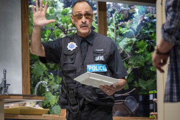 Poliisi, jolla on aurinkolasit ja liivit, joissa lukee POLIISI, nostaa yhtä kättä ja pitää tablettia kädessään keittiössä. Hänen takanaan olevasta ikkunasta näkyy puita ja vehreyttä.