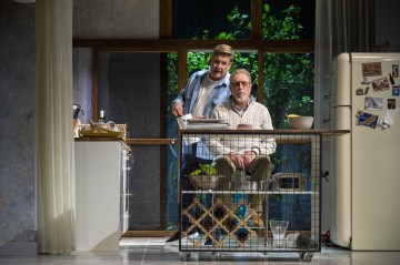 Kaksi miestä keittiössä; toinen istuu pöydän ääressä ja näyttää vakavalta, kun toinen seisoo hänen takanaan ja nojaa kädellään hänen olkapäälleen. Taustalla näkyy jääkaappi, jossa on magneetteja ja valokuvia, kasveja ja ikkuna, jossa on viherkasveja.