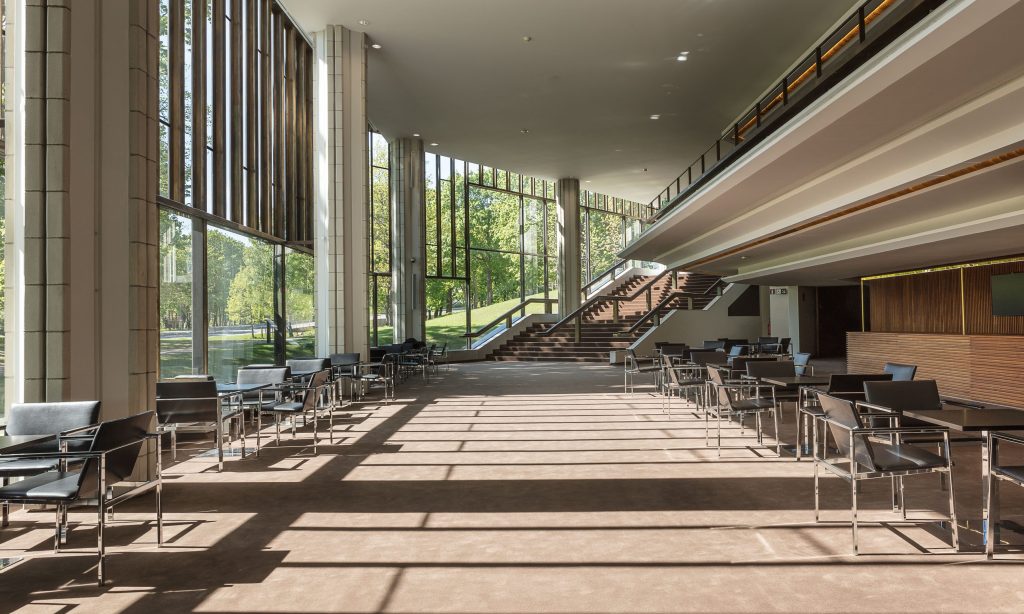 Tilava, moderni aula, jossa on lattiasta kattoon ulottuvat ikkunat, metalli- ja nahkatuoleja pienissä pöydissä, auringonvalo paistaa läpi, puuportaat ja ulkona näkyvät vihreät puut.