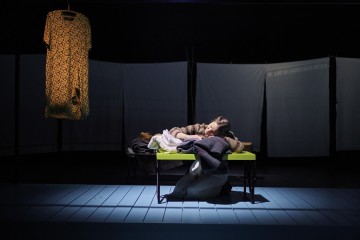 Henkilö makaa vihreällä penkillä, pää käsien varassa, vaatteiden ympäröimänä. Heidän yläpuolellaan roikkuu kuviollinen mekko, ja valkoiset paneelit muodostavat taustan hämärästi valaistulla näyttämöllä.