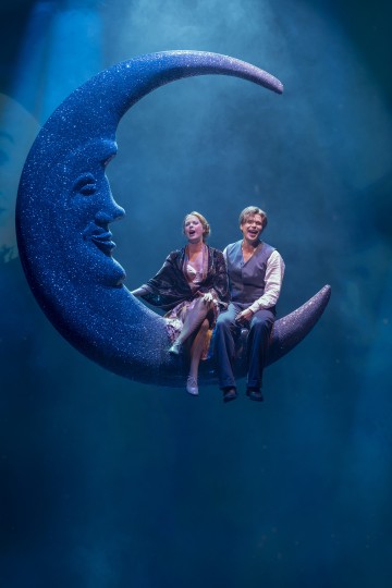 Kaksi esiintyjää vintage-tyylisissä puvuissa istuu suuren, kuohuvan kuunsirpin ja kasvot sisältävän rekvisiitan päällä, joka on ripustettu siniseen näyttämövalaistukseen ja luo unenomaisen, teatraalisen kohtauksen.