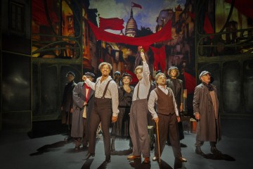 Ryhmä näyttelijöitä poseeraa näyttämöllä dramaattisesti aikakauden puvuissa, pitelee punaisia lippuja kädessään vanhojen pariisilaisten rakennusten taustalla ja heiluttaa banderolleja, mikä tuo mieleen vallankumouskohtauksen.