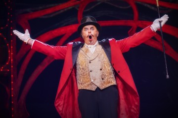 Sirkusjohtaja, jolla on punainen takki, kultainen liivi, valkoiset hansikkaat ja musta silinterihattu, seisoo kädet ojennettuina ja suu auki ja näyttää ilmoittavan tai toivottavan yleisön tervetulleeksi.