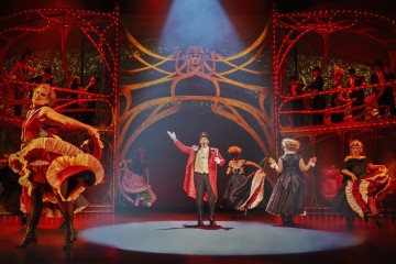 Teatterin näyttämökohtaus, jossa punatakkinen sirkustirehtööri seisoo keskellä näyttämöä valonheittimen alla, häntä ympäröivät värikkäisiin can-can-pukuihin pukeutuneet tanssijat, esiintyjät parvekkeilla ja koristeellinen punainen ja kultainen koristelu.