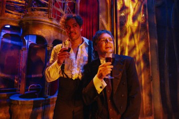 Kaksi viralliseen, vintage-tyyliseen pukeutumiseen pukeutunutta miestä pitelee punaviinilaseja ja seisoo lähekkäin lämpimästi valaistussa, koristeellisessa huoneessa, jossa on kullan ja punaisen värinen sisustus, joka tuo mieleen teatterin tai historiallisen miljöön.