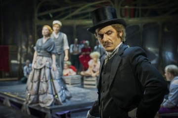 Etualalla seisoo mustaan silinterihattuun ja viktoriaaniseen pukuun pukeutunut mies, joka näyttää vakavalta. Hänen takanaan on näyttelijöitä, joilla on hienostuneet aikakauden vaatteet ja jotka esiintyvät teatterilavalla, jossa on dramaattinen valaistus.