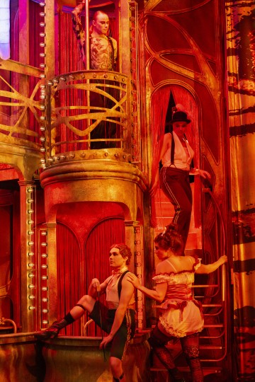 Neljä esiintyjää kabaree-tyylisissä puvuissa poseeraavat koristeellisella, kullanpunaisella näyttämöllä, jossa on parvekkeita ja kaarevia metalliosia ja joka on valaistu lämpimässä, dramaattisessa valaistuksessa.