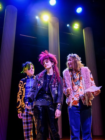 Kolme ihmistä värikkäissä, punk-vaikutteisissa asuissa seisoo lavalla kirkkaiden valojen ja korkeiden pylväiden alla ja näyttää ilmeikkäältä ja sitoutuneelta kuin teatterituotannossa.
