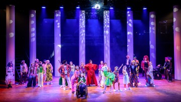Värikkäisiin, eklektisiin pukuihin pukeutunut esiintyjäjoukko seisoo lavalla, jossa on korkeat valkoiset pylväät ja dramaattiset valonheittimet, ja luo eloisan teatterikohtauksen valaistun diskopallon alla.