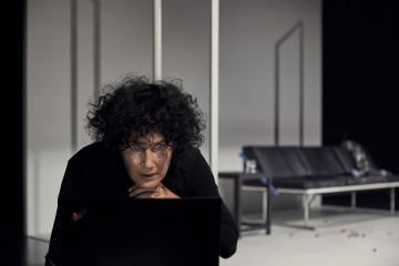 Mustiin pukeutunut tummahiuksinen henkilö, jolla on kiharat tummat hiukset, nojaa eteenpäin ja katsoo keskittyneesti kannettavaa tietokonetta minimalistisessa huoneessa, jonka taustalla on musta sohva ja geometrisia valkoisia rakenteita.