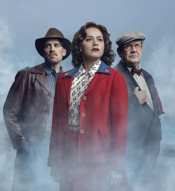 Kolme ihmistä seisoo sumussa: edessä on nainen, jolla on punainen takki ja kuviollinen pusero, vasemmalla mies, jolla on nahkatakki ja hattu, ja oikealla vanhempi mies, jolla on litteä lakki, silmälasit ja rusetti.