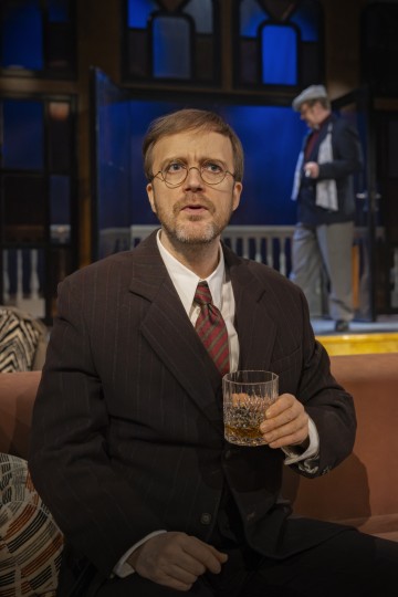 Sohvalla istuu mies, jolla on puikkopuku ja silmälasit, viskilasi kädessään ja katsoo mietteliäästi ylöspäin. Toinen mies seisoo taustalla oven vieressä hämärässä huoneessa, jossa on sininen valaistus.