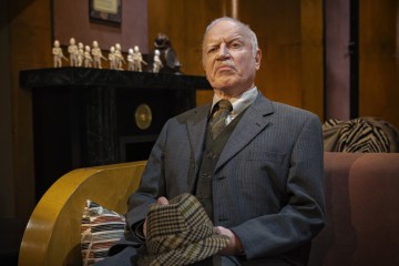 Harmaapukuinen vanhempi mies istuu sohvalla ja pitää käsissään ruudullista hattua. Hän näyttää vakavalta ja mietteliäältä. Hänen takanaan on musta takkatuli, jossa on pieniä patsaita, ja puuverhoiltu seinä.