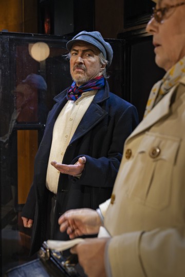 Vanhempi mies, jolla on litteä lakki ja tumma takki, seisoo toinen käsi kohotettuna ja katsoo toista beigeen takkiin pukeutunutta henkilöä, jolla on kirjoituskone kädessään. He näyttävät keskustelevan vakavasti sisätiloissa.