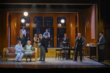 Ryhmä näyttelijöitä esiintyy kirkkaasti valaistulla näyttämöllä, joka muistuttaa olohuoneen lavastusta, ja ympärillä on sekä virallisesti että rennosti pukeutuneita ihmisiä, joista osa istuu, osa seisoo ja osa keskustelee vilkkaasti.