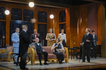 Seitsemän viralliseen tai puoliviralliseen asuun pukeutunutta henkilöä on lavastettu tyylikästä olohuonetta muistuttavaan lavasteeseen. Jotkut seisovat, toiset istuvat sohvalla, ja kaikki näyttävät olevan mukana vakavassa keskustelussa tai dramaattisessa kohtauksessa.