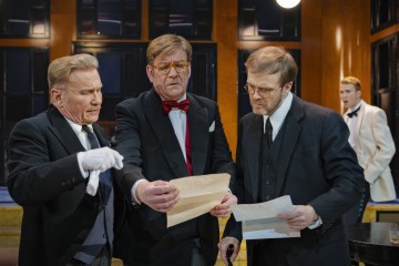 Kolme muodollisiin pukuihin pukeutunutta miestä tutkii tiiviisti asiakirjoja yhdessä, ja he vaikuttavat vakavilta ja keskittyneiltä. Toinen mies valkoisessa smokissa seisoo taustalla ja tarkkailee heitä hyvin valaistussa huoneessa, jossa on suuret ikkunat.