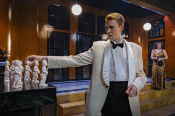 Valkoiseen smokkiin pukeutunut mies kurottaa nappulaa shakkilaudalta, jossa on figuurinappuloita, kun taas kultamustaan mekkoon pukeutunut nainen seisoo taustalla portailla ja katsoo häntä. Ympäristö näyttää olevan tyylikäs, moderni huone.