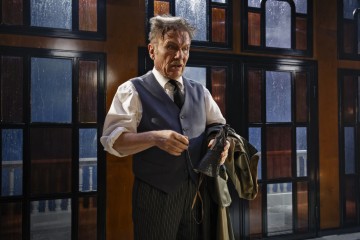 Vanhempi mies, jolla on epäsiisti tukka ja liivi, seisoo yöllä sisätiloissa takki ja sateenvarjo kädessään, ja hänen takanaan on suuret ikkunat, joista näkyy sade ulkona.