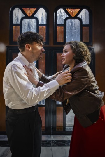 Valkoiseen paitaan pukeutunut mies ja ruskeaan takkiin ja punaiseen hameeseen pukeutunut nainen seisovat lähekkäin, toisiaan syleillen, ahdistuneen ja tunteikkaan näköisinä lasimaalausten edessä.