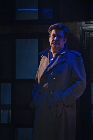 Mies seisoo hämärässä, tunnelmallisessa valaistuksessa trenssitakki päällään, kädet taskuissa, ja katsoo eteenpäin vakavalla ilmeellä. Tausta on tumma ja siinä on sinisiä korostuksia.