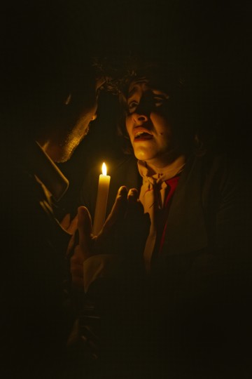 Kahta pimeässä olevaa ihmistä valaisee yksi kynttilä, jota toinen heistä pitää kädessään. Valo korostaa heidän kasvojaan, luo dramaattisia varjoja ja antaa kohtaukselle jännittyneen, tunteikkaan tunnelman.